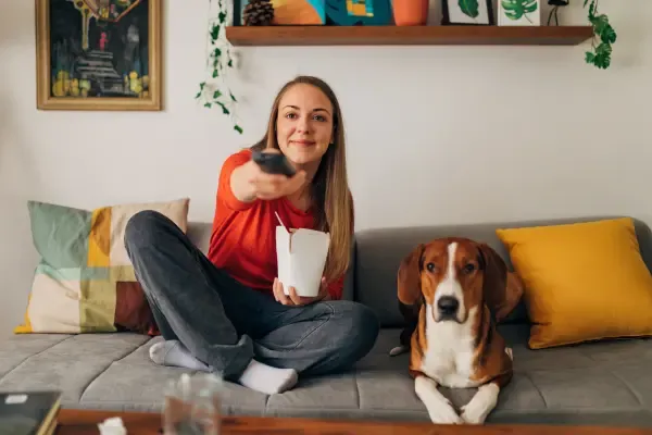 A woman sits on a sofa with her dog, holding a takeout box and pointing a TV remote.